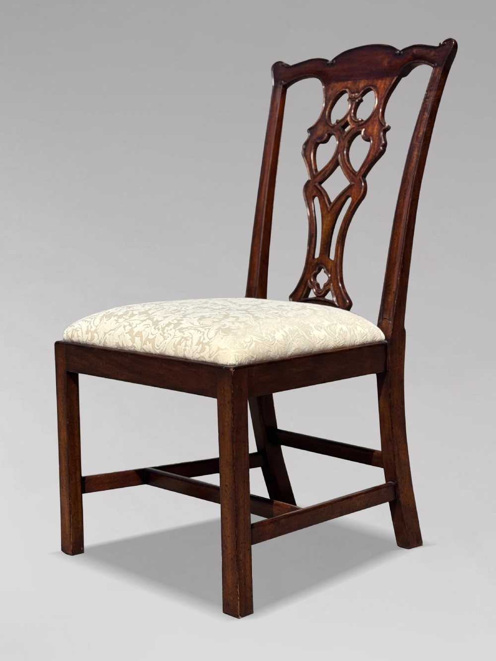 A set of six Edwardian mahogany dining chairs featuring pierced carved back splats, serpentine top rails, cream upholstered seats, and straight square legs with H-stretchers. View IX