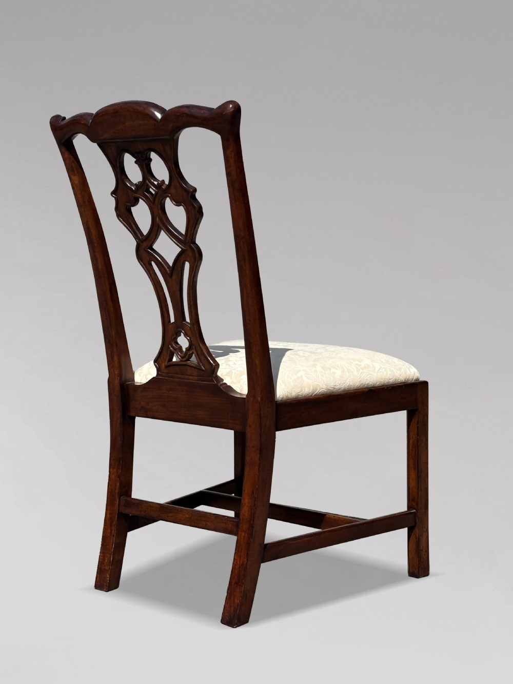 A set of six Edwardian mahogany dining chairs featuring pierced carved back splats, serpentine top rails, cream upholstered seats, and straight square legs with H-stretchers. View X