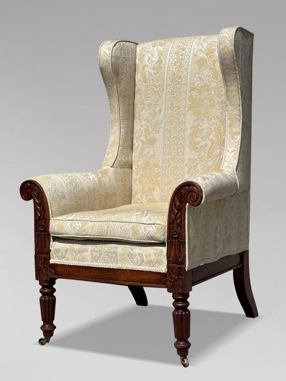A 19th-century William IV wingback armchair upholstered in cream patterned damask fabric with carved mahogany arm supports and turned front legs on brass castors.