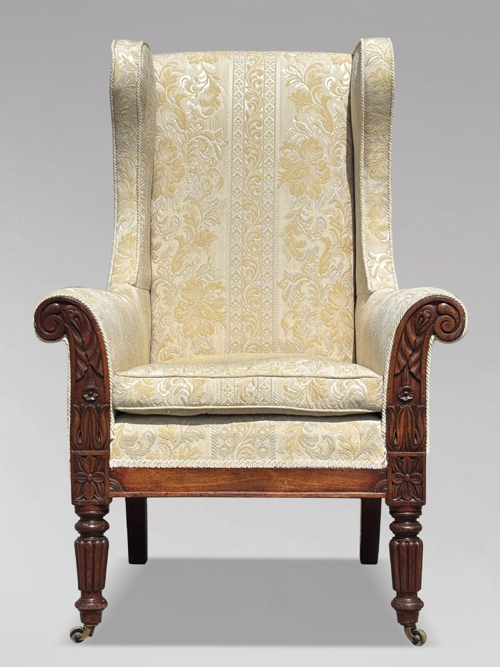 A 19th-century William IV wingback armchair upholstered in cream patterned damask fabric with carved mahogany arm supports and turned front legs on brass castors. View II