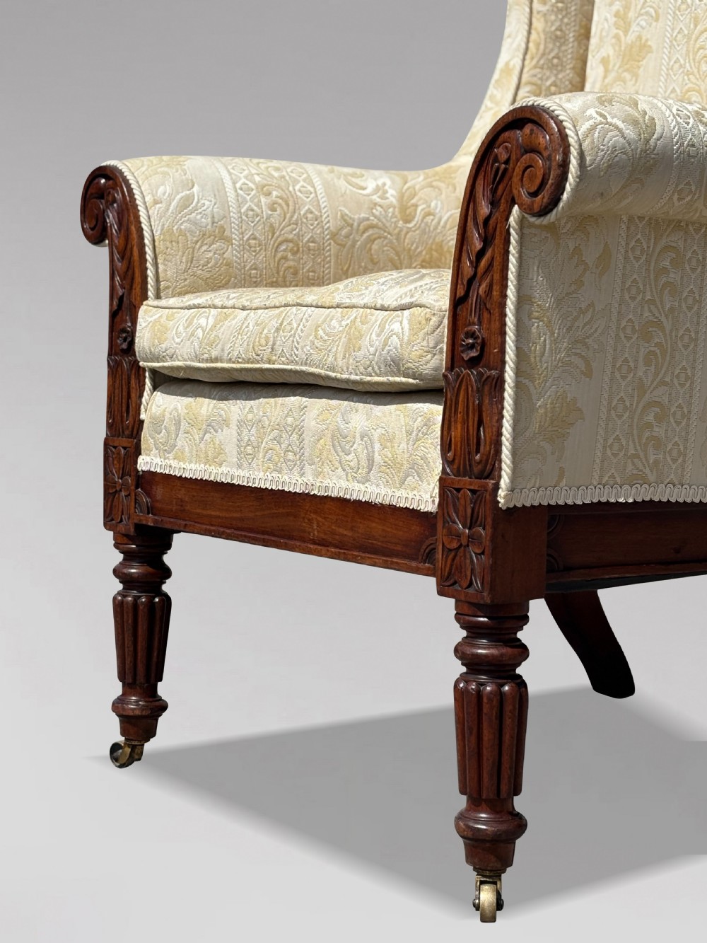 A 19th-century William IV wingback armchair upholstered in cream patterned damask fabric with carved mahogany arm supports and turned front legs on brass castors. View III