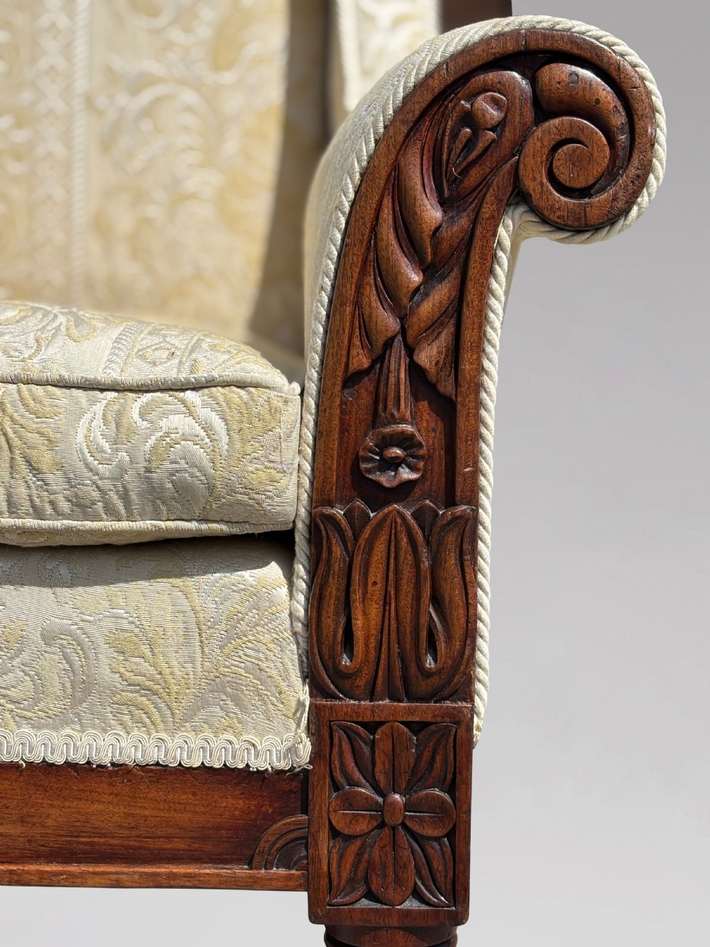 A 19th-century William IV wingback armchair upholstered in cream patterned damask fabric with carved mahogany arm supports and turned front legs on brass castors. View IV