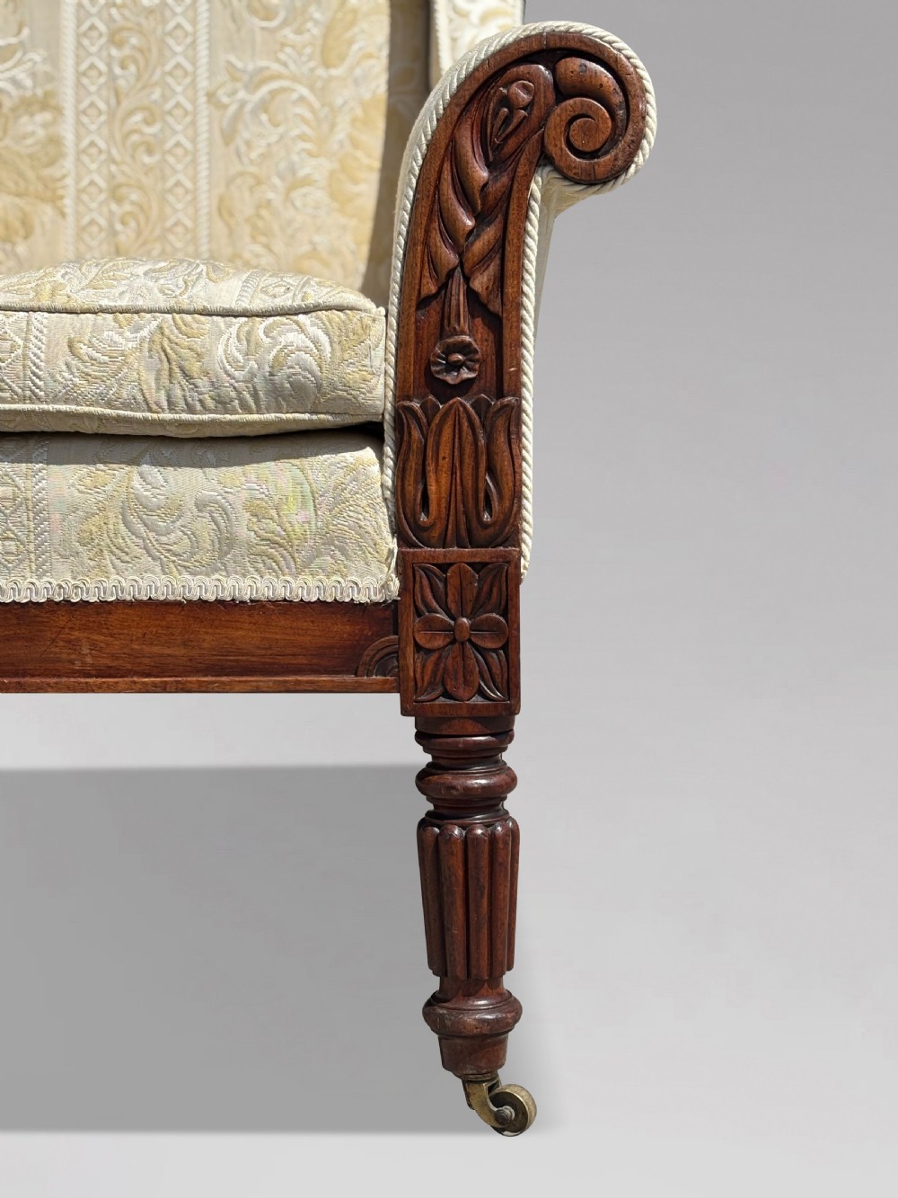 A 19th-century William IV wingback armchair upholstered in cream patterned damask fabric with carved mahogany arm supports and turned front legs on brass castors. View V
