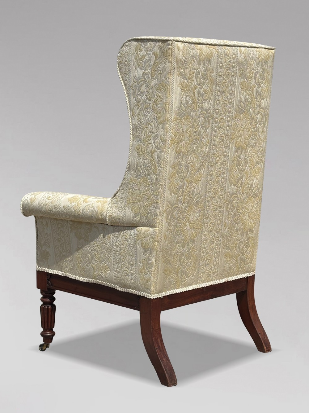 A 19th-century William IV wingback armchair upholstered in cream patterned damask fabric with carved mahogany arm supports and turned front legs on brass castors. View VI