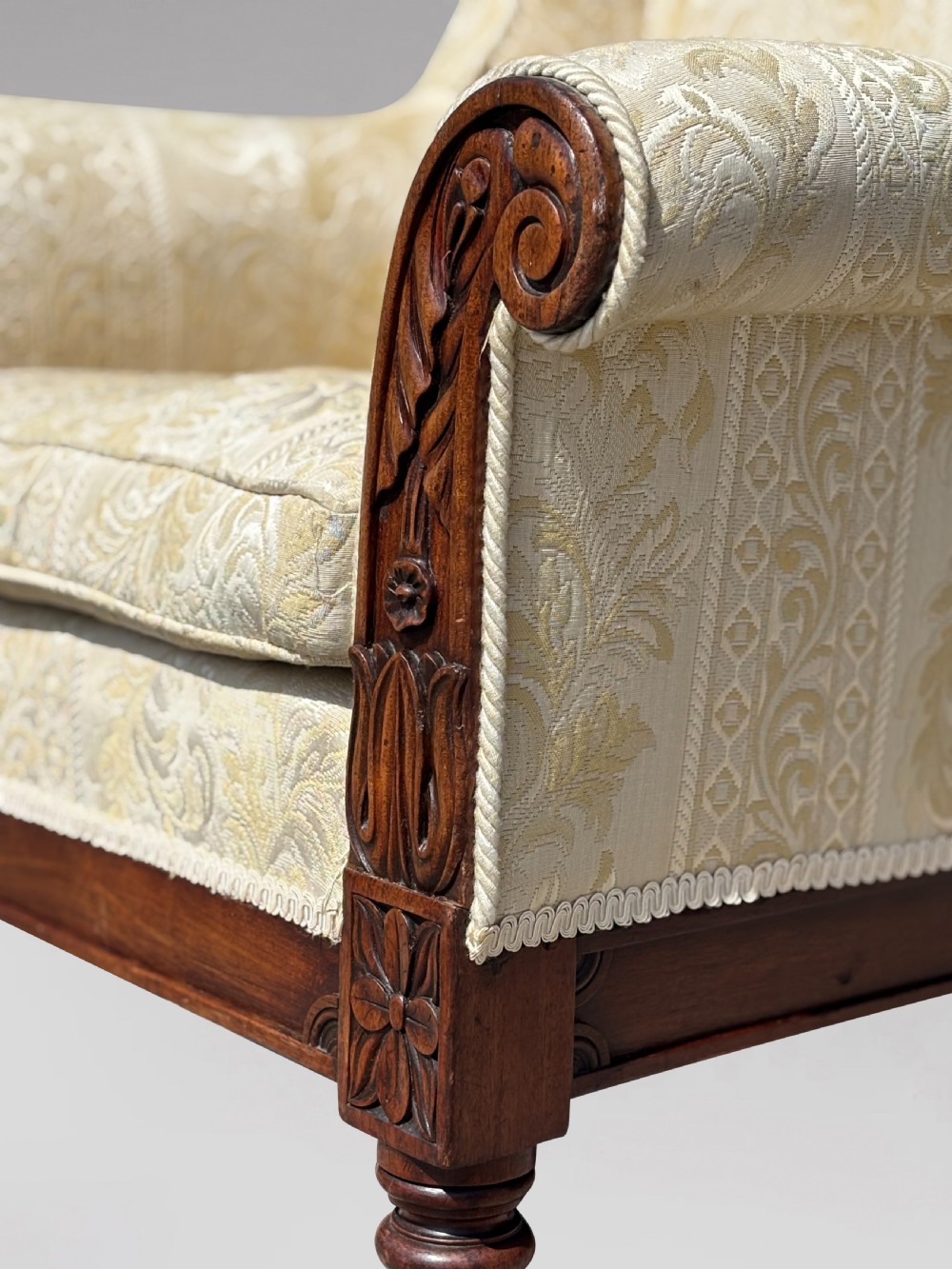 A 19th-century William IV wingback armchair upholstered in cream patterned damask fabric with carved mahogany arm supports and turned front legs on brass castors. View VII