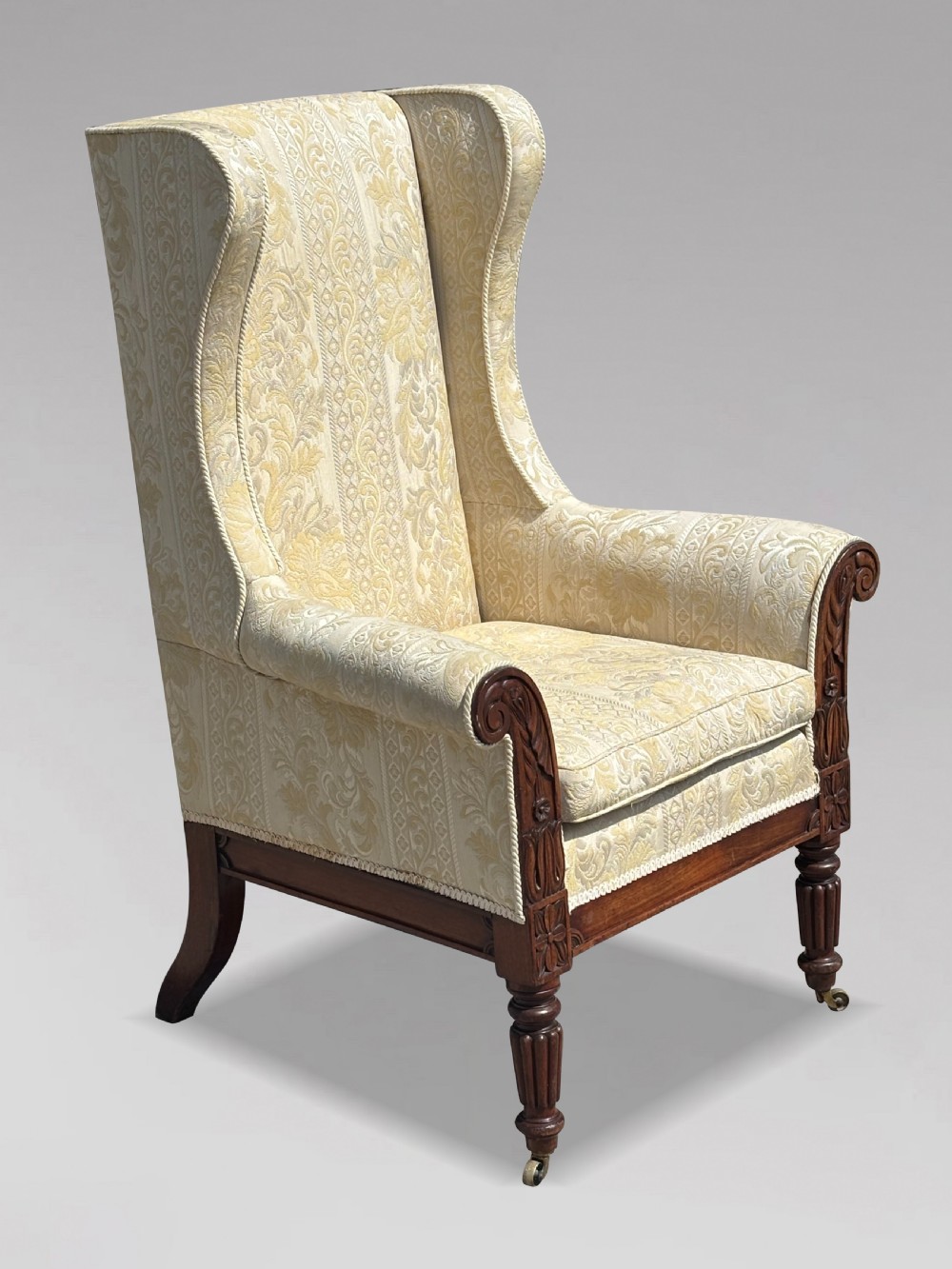 A 19th-century William IV wingback armchair upholstered in cream patterned damask fabric with carved mahogany arm supports and turned front legs on brass castors. View VIII