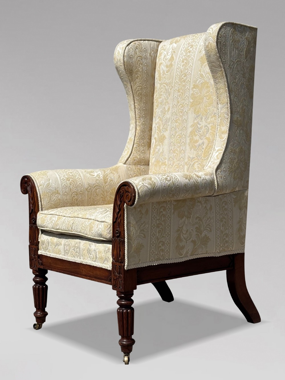 A 19th-century William IV wingback armchair upholstered in cream patterned damask fabric with carved mahogany arm supports and turned front legs on brass castors. View IX