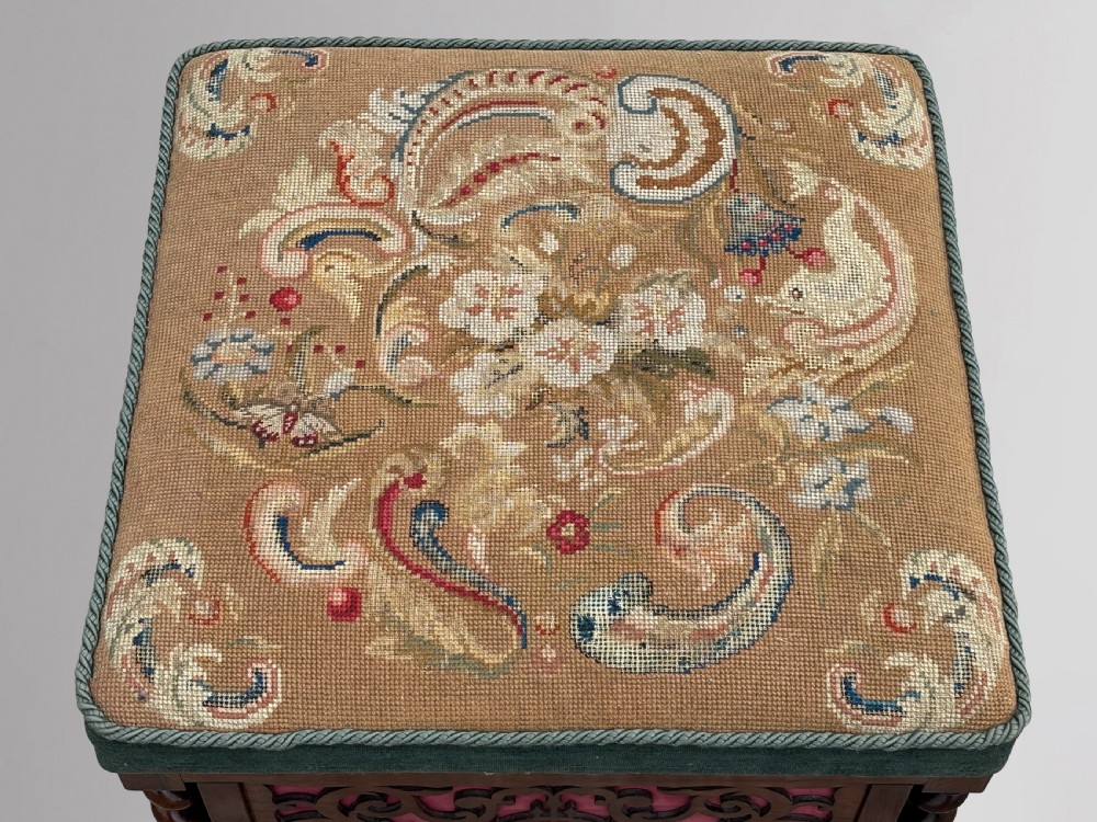 A Victorian rosewood piano stool featuring a square tapestry-upholstered top with green velvet edging, carved fretwork side panels, corner barley twist columns, and bun feet. View IV