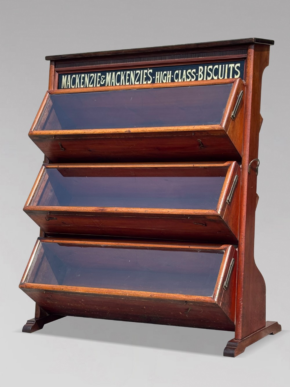 A late 19th-century mahogany three-tiered biscuit display cabinet featuring angled glass fronts, brass handles, and a painted advertising header for Mackenzies Biscuits.
