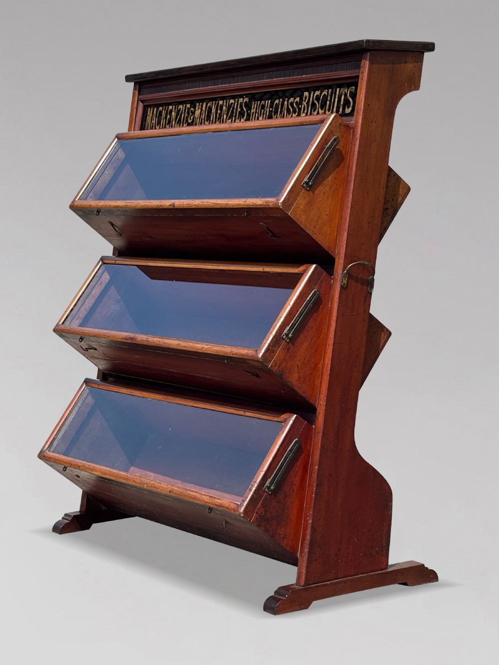 A late 19th-century mahogany three-tiered biscuit display cabinet featuring angled glass fronts, brass handles, and a painted advertising header for Mackenzies Biscuits. View II