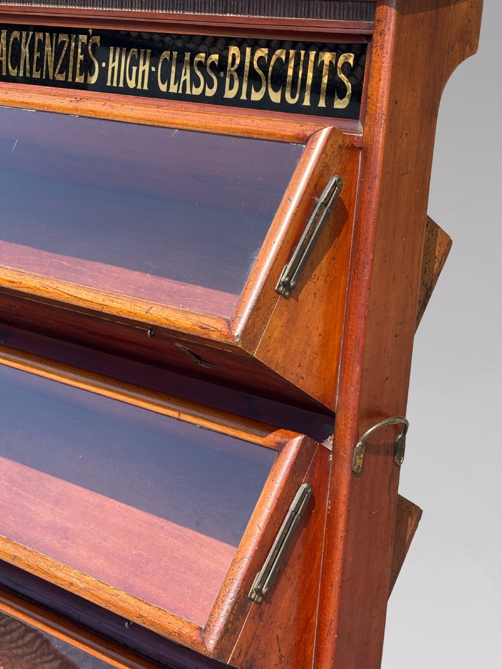 A late 19th-century mahogany three-tiered biscuit display cabinet featuring angled glass fronts, brass handles, and a painted advertising header for Mackenzies Biscuits. View VI