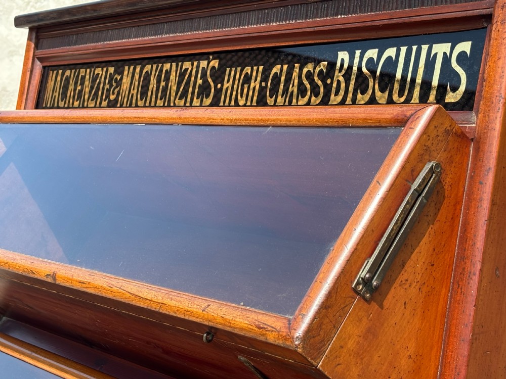 A late 19th-century mahogany three-tiered biscuit display cabinet featuring angled glass fronts, brass handles, and a painted advertising header for Mackenzies Biscuits. View X