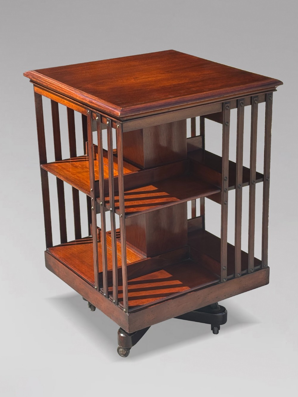 A late 19th-century Victorian mahogany revolving bookcase featuring three tiers of open shelving with vertical side slats and a square top, resting on castors.