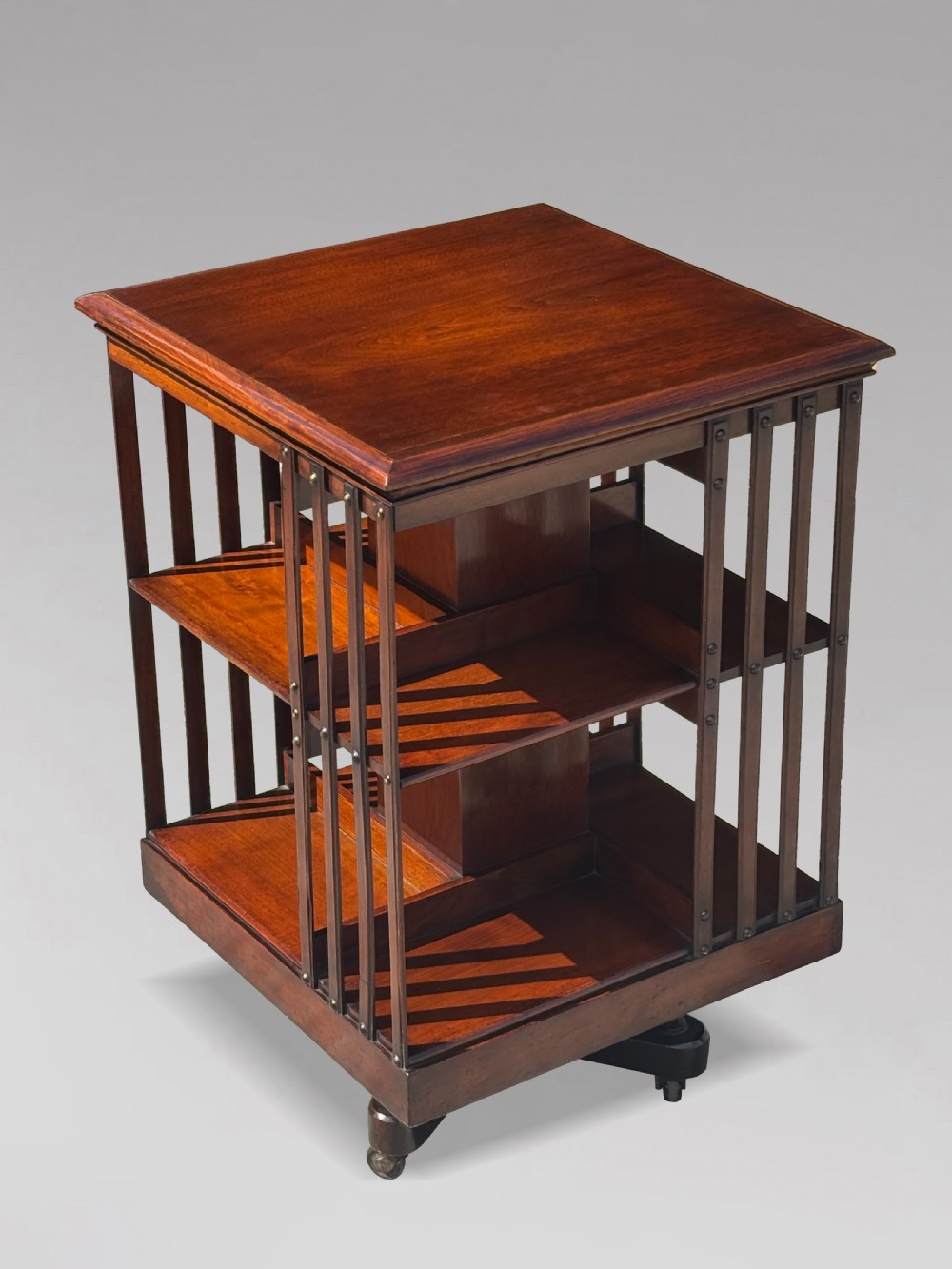 A late 19th-century Victorian mahogany revolving bookcase featuring three tiers of open shelving with vertical side slats and a square top, resting on castors. View II
