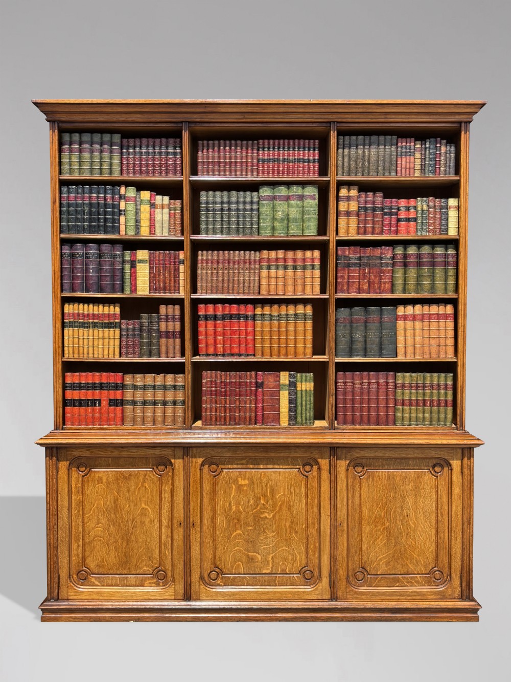 A tall 19th-century French oak open bookcase featuring a three-bay upper shelving section filled with books over a triple-door panelled cupboard base.