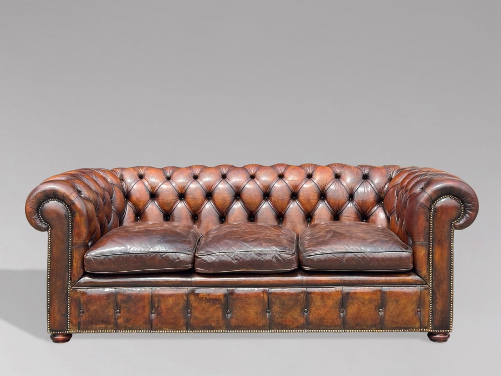 A nineteenth-century three-seater Chesterfield sofa upholstered in deep buttoned chestnut brown leather with brass stud detailing along the rolled arms. View X