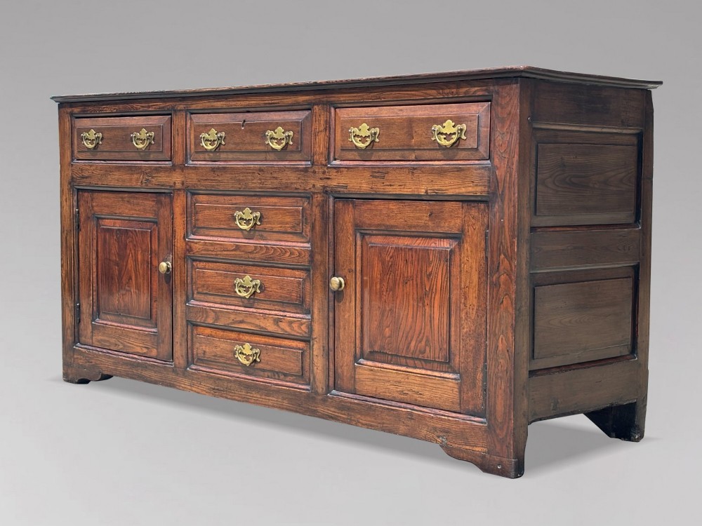 An 18th-century George II solid oak dresser base featuring multiple drawers, panelled cupboard doors, and brass drop handles.