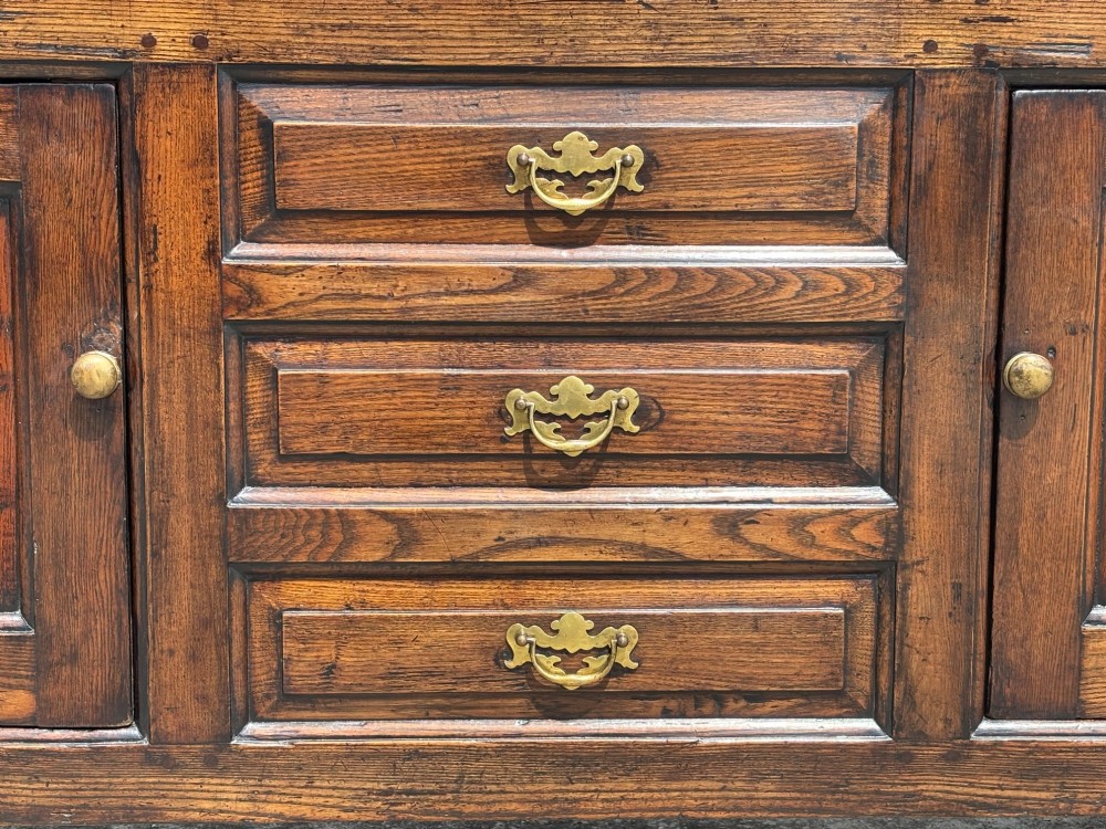 An 18th-century George II solid oak dresser base featuring multiple drawers, panelled cupboard doors, and brass drop handles. View V