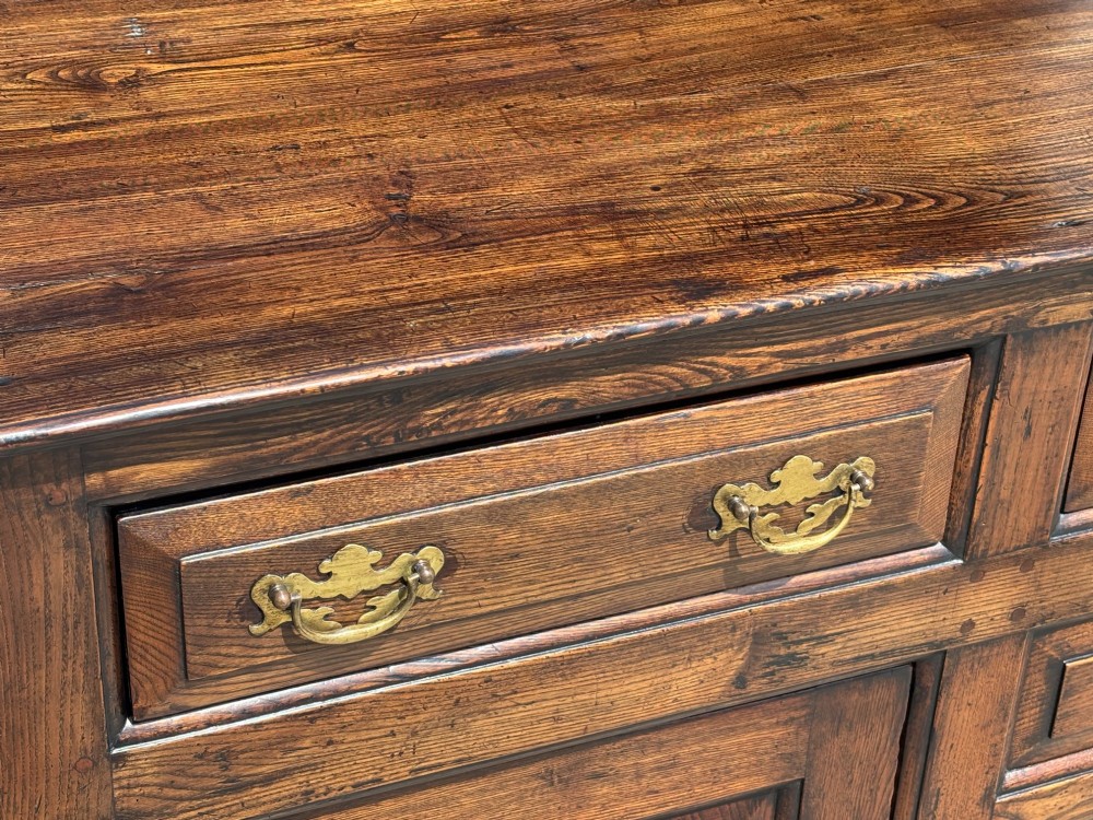 An 18th-century George II solid oak dresser base featuring multiple drawers, panelled cupboard doors, and brass drop handles. View VI
