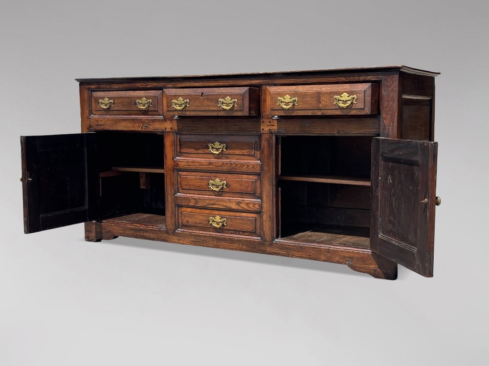An 18th-century George II solid oak dresser base featuring multiple drawers, panelled cupboard doors, and brass drop handles. View XII