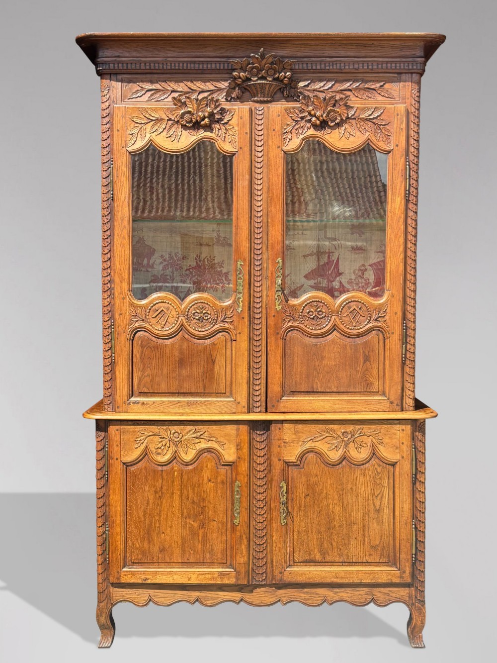 An 18th-century carved oak French buffet deux corps featuring upper glazed doors lined with fabric above a panelled lower cabinet with relief carvings.