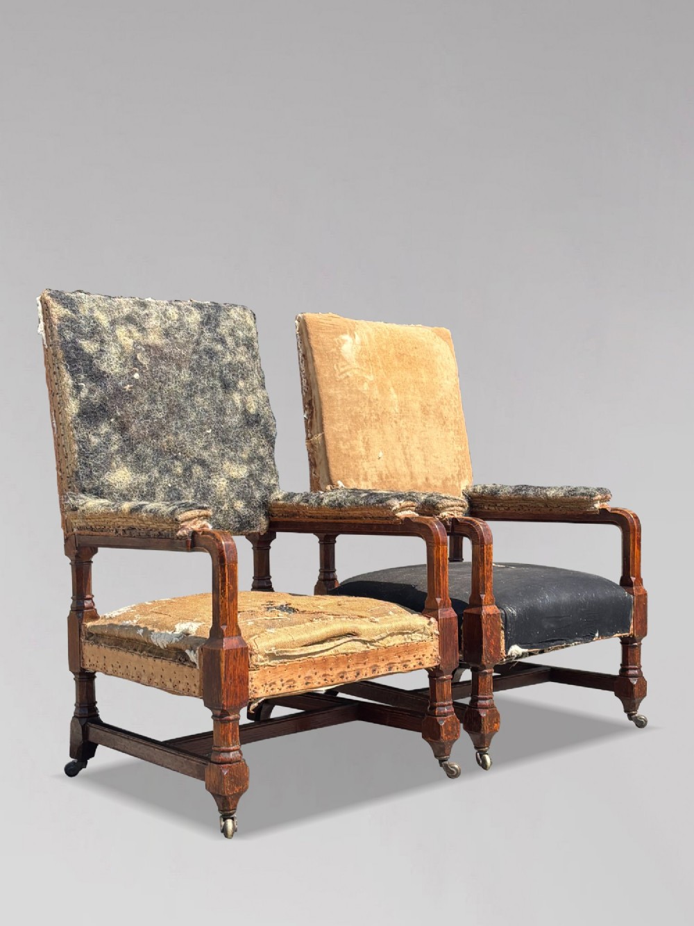 A pair of 19th-century Victorian oak Gothic Revival open armchairs featuring carved timber frames, turned legs with brass castors, and exposed upholstery padding.