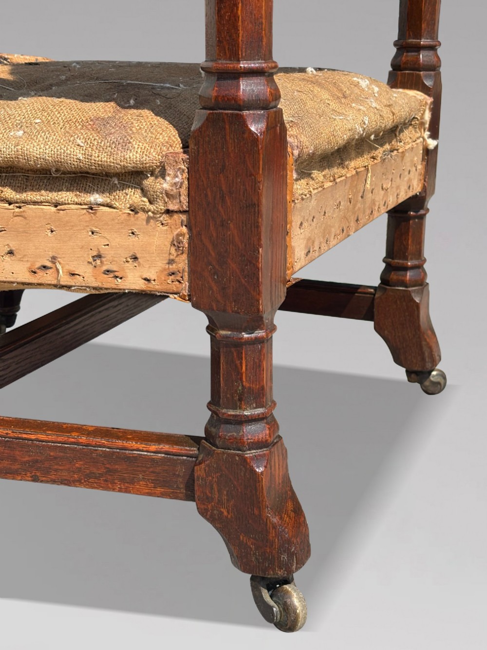 A pair of 19th-century Victorian oak Gothic Revival open armchairs featuring carved timber frames, turned legs with brass castors, and exposed upholstery padding. View X