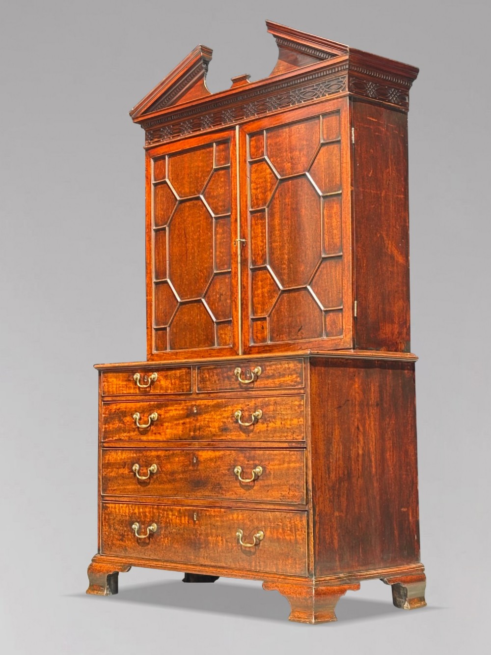 An 18th-century George III mahogany bookcase on chest featuring a broken pediment, carved frieze, geometric astragal glazed doors, and four lower drawers with brass handles.