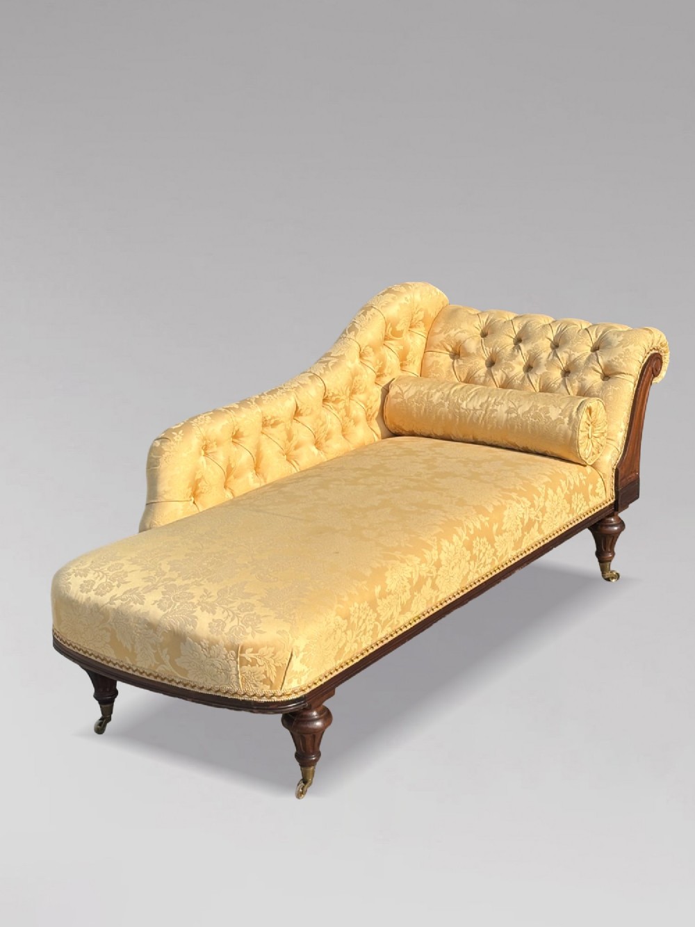 A 19th-century Regency mahogany chaise longue upholstered in yellow damask fabric with deep buttoning, a bolster cushion, and turned legs on brass castors. View VIII