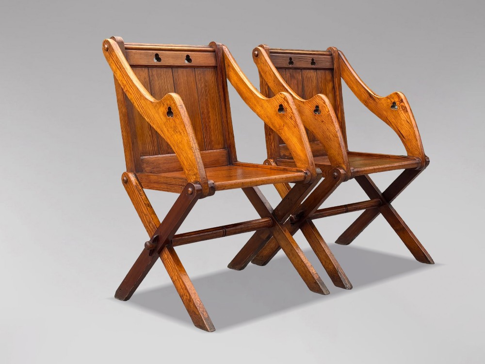 A pair of Victorian pitch pine Glastonbury armchairs featuring sloped armrests, X-frame bases, and pierced trefoil motifs on the back panels.