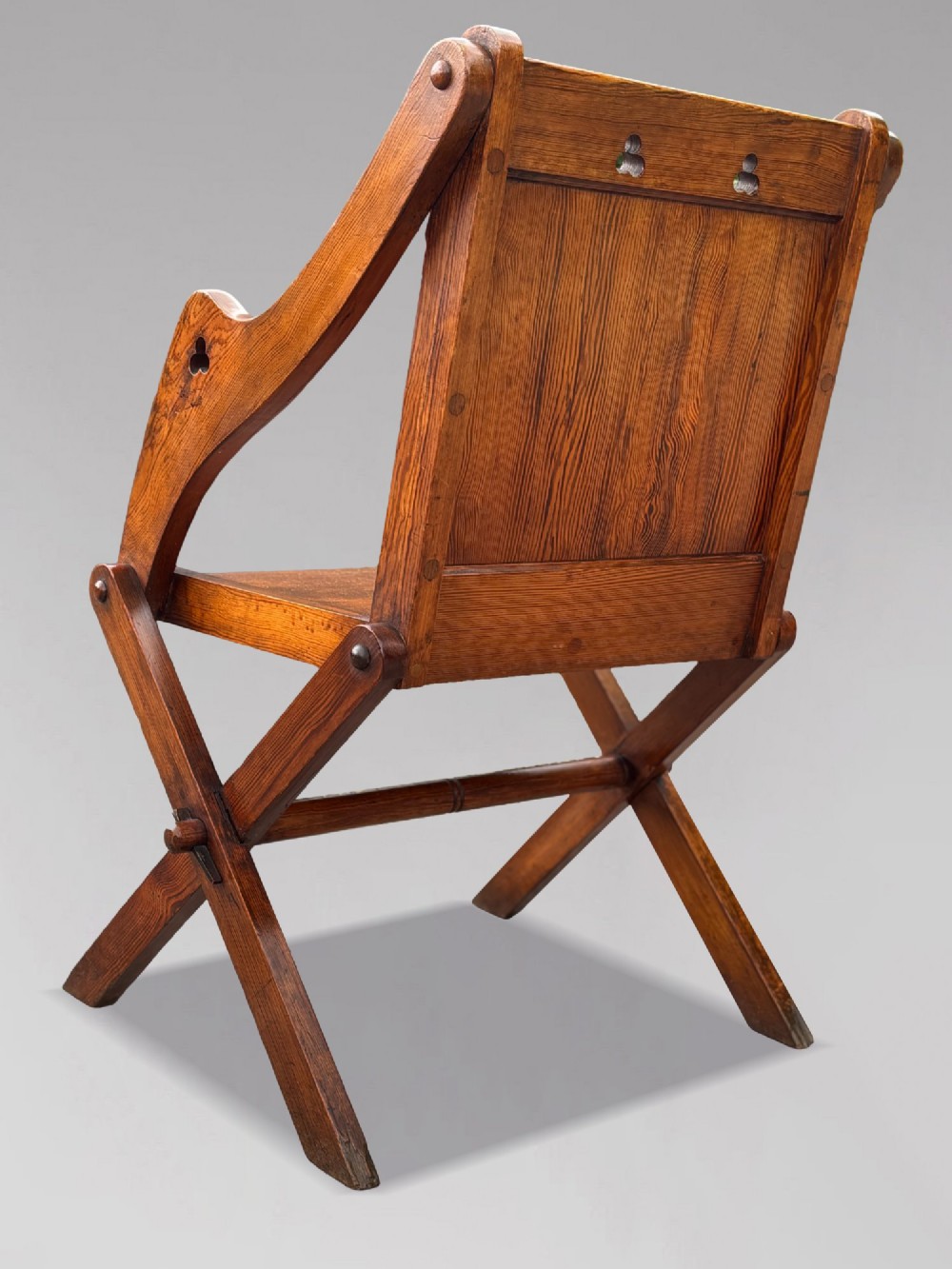 A pair of Victorian pitch pine Glastonbury armchairs featuring sloped armrests, X-frame bases, and pierced trefoil motifs on the back panels. View IV