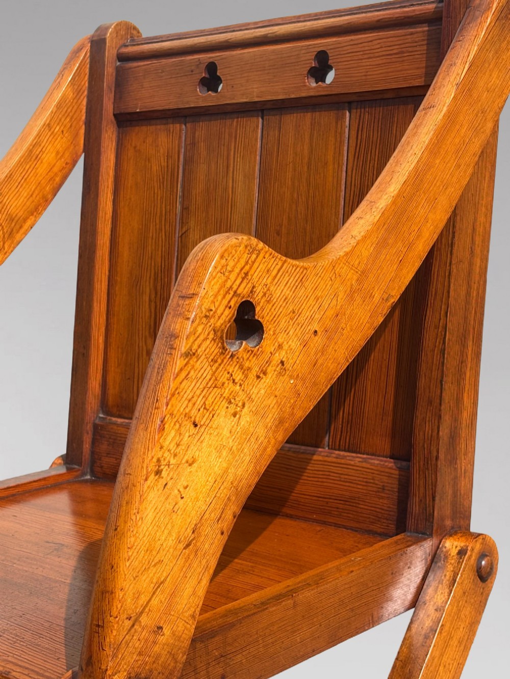 A pair of Victorian pitch pine Glastonbury armchairs featuring sloped armrests, X-frame bases, and pierced trefoil motifs on the back panels. View V