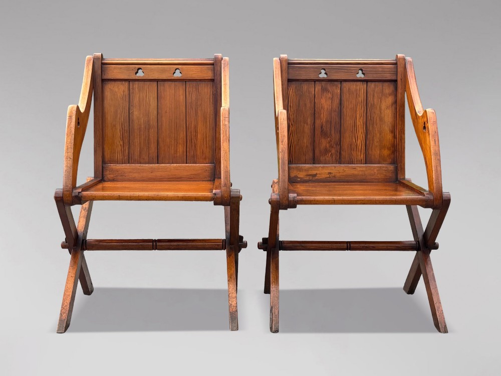 A pair of Victorian pitch pine Glastonbury armchairs featuring sloped armrests, X-frame bases, and pierced trefoil motifs on the back panels. View X