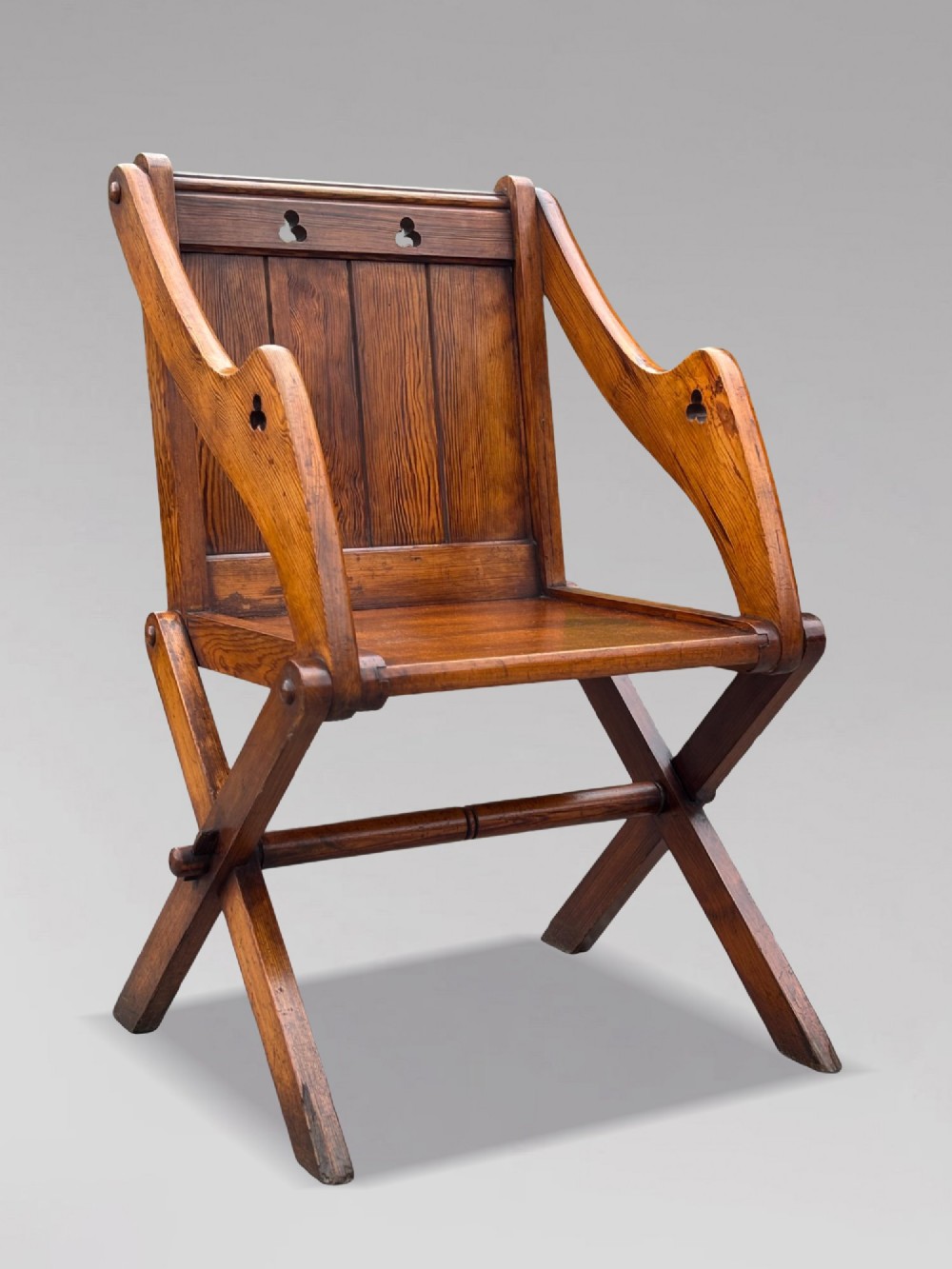 A pair of Victorian pitch pine Glastonbury armchairs featuring sloped armrests, X-frame bases, and pierced trefoil motifs on the back panels. View XII