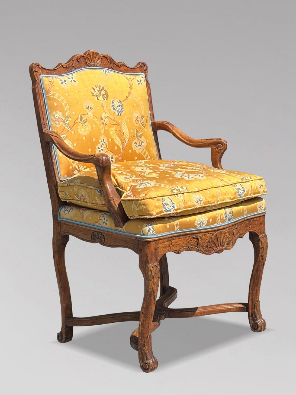An 18th-century French carved walnut armchair with cabriole legs, an X-form stretcher, and yellow floral upholstered cushions finished with blue trim.