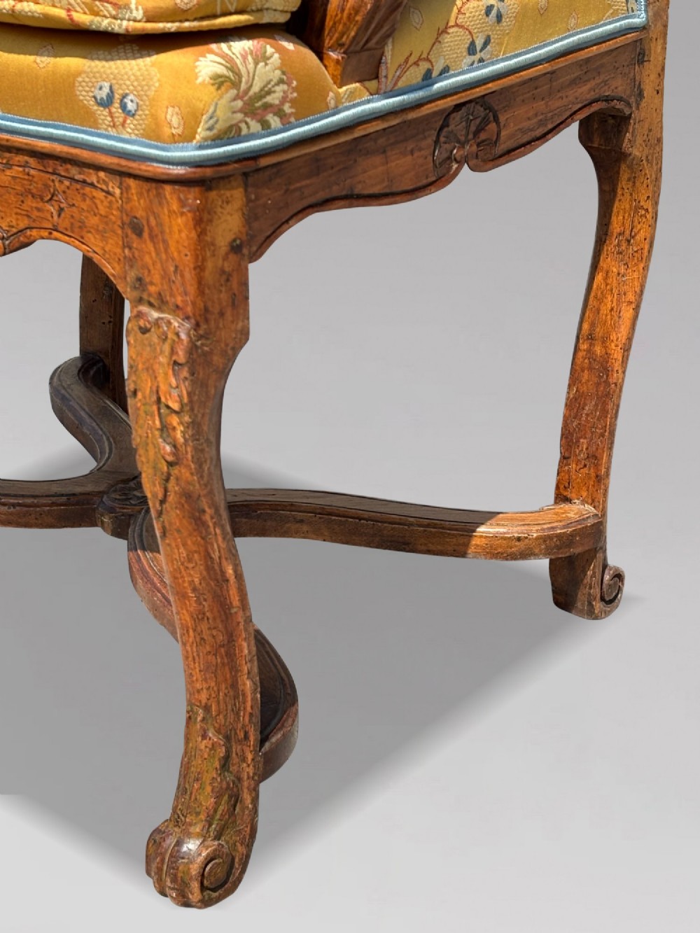 An 18th-century French carved walnut armchair with cabriole legs, an X-form stretcher, and yellow floral upholstered cushions finished with blue trim. View X