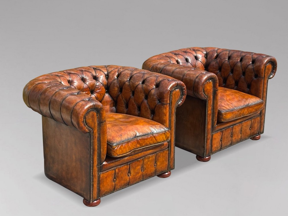 A pair of late Victorian deep-buttoned brown leather Chesterfield club armchairs with rolled arms, brass studding, and turned wooden feet. View II