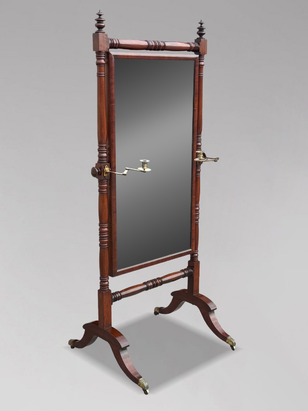 An early 19th-century Regency mahogany cheval mirror featuring turned uprights, brass candle sconces, finials, and splayed legs on brass castors.