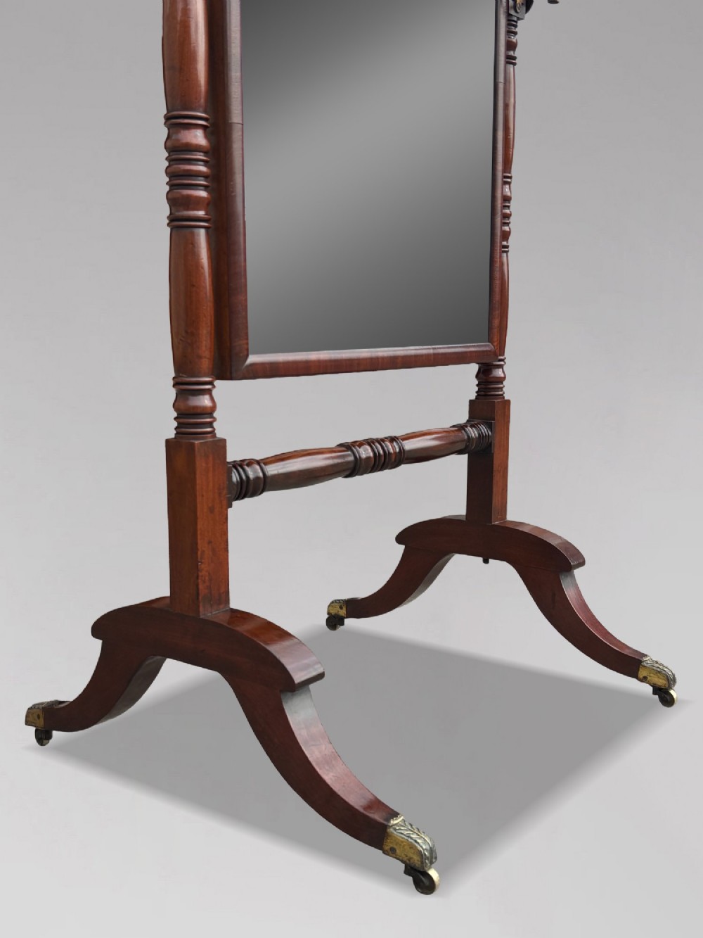 An early 19th-century Regency mahogany cheval mirror featuring turned uprights, brass candle sconces, finials, and splayed legs on brass castors. View VI