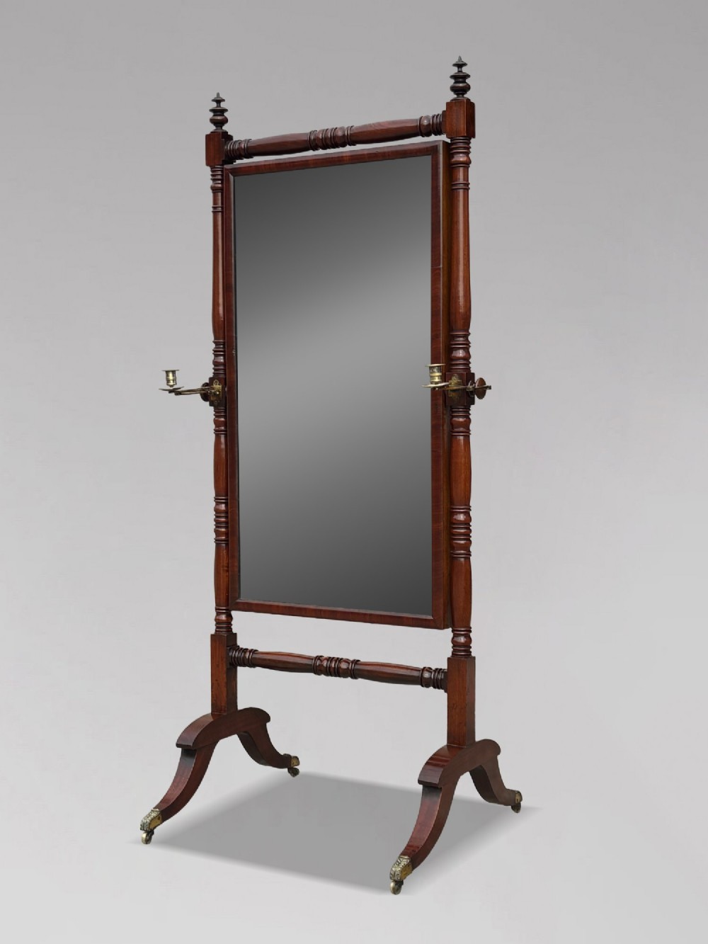 An early 19th-century Regency mahogany cheval mirror featuring turned uprights, brass candle sconces, finials, and splayed legs on brass castors. View X