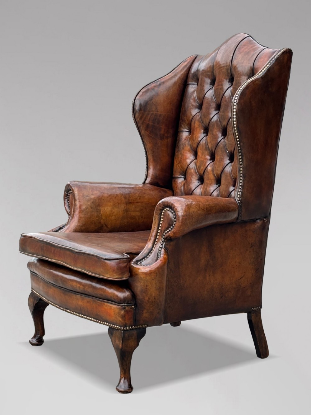 A 19th-century Victorian wingback armchair upholstered in deep-buttoned warm brown leather with brass stud detailing and carved mahogany legs. View V