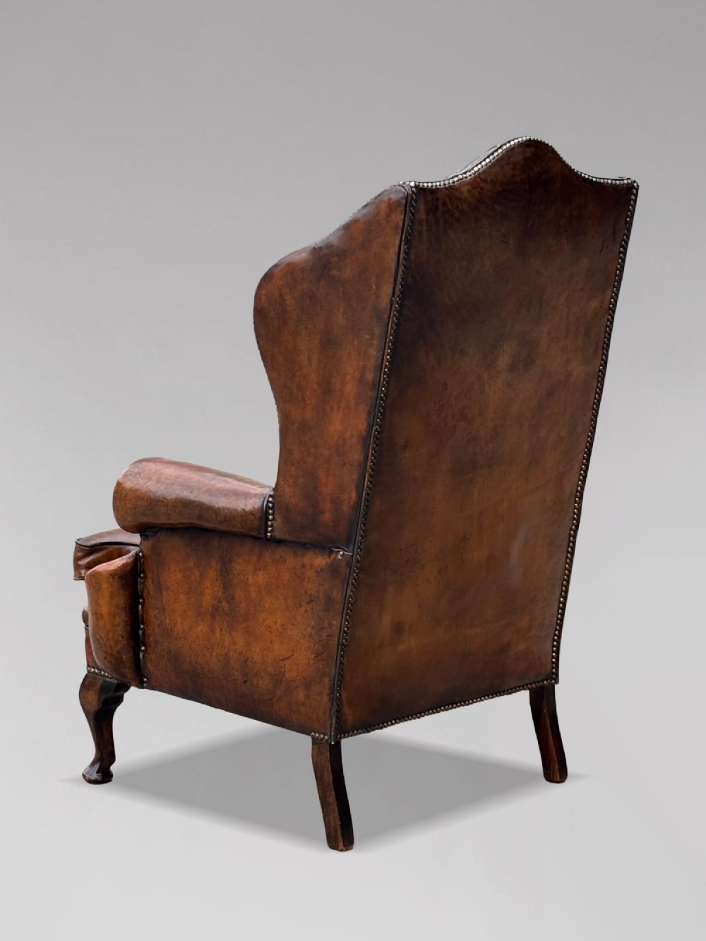 A 19th-century Victorian wingback armchair upholstered in deep-buttoned warm brown leather with brass stud detailing and carved mahogany legs. View VI