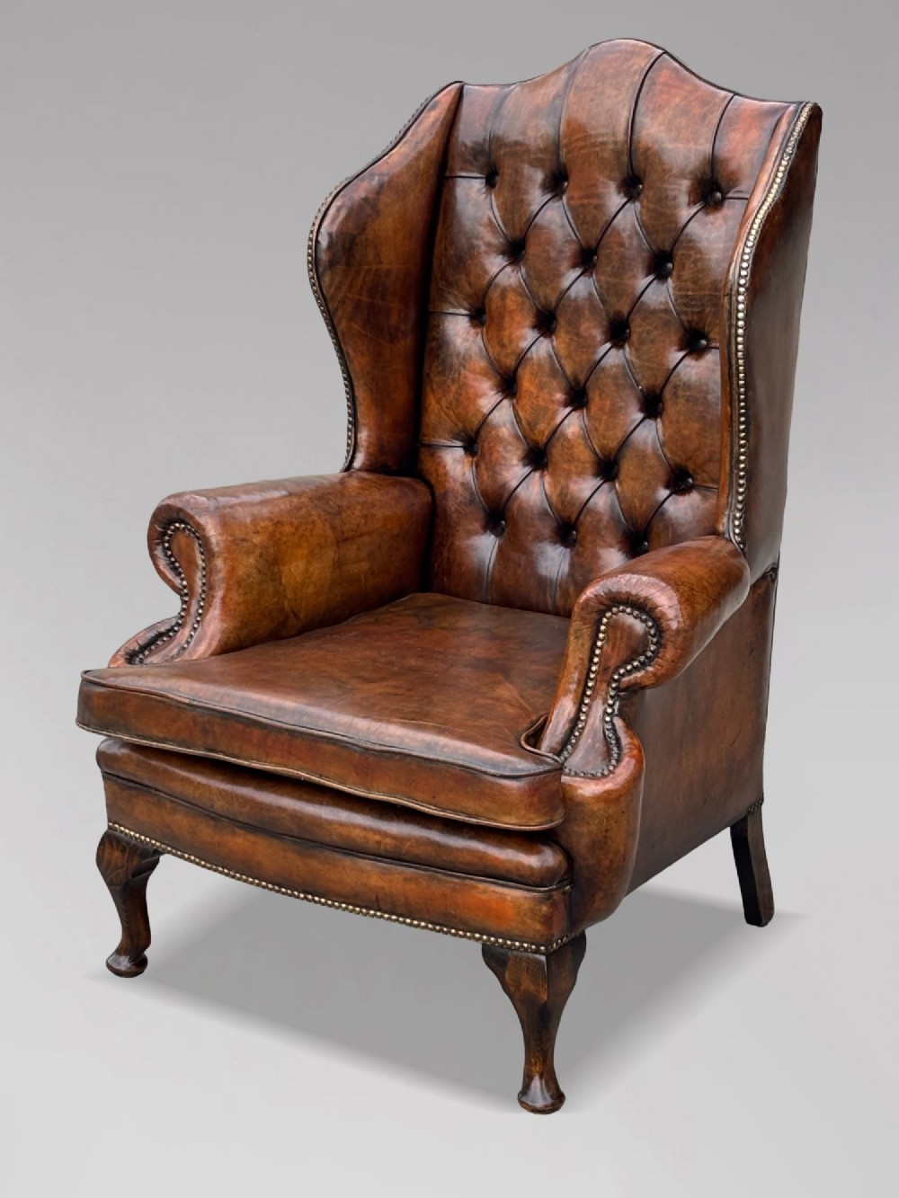 A 19th-century Victorian wingback armchair upholstered in deep-buttoned warm brown leather with brass stud detailing and carved mahogany legs. View VII
