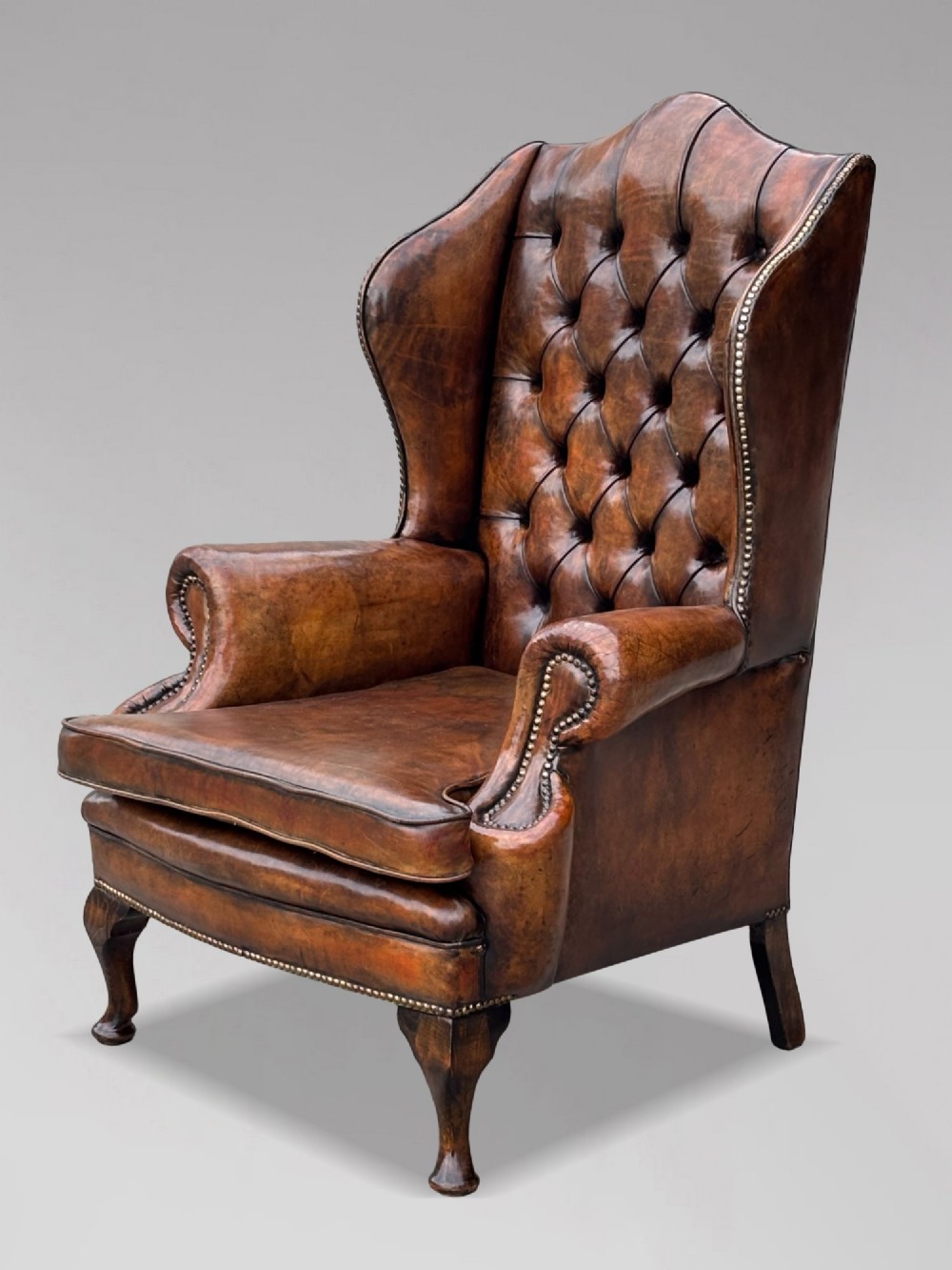 A 19th-century Victorian wingback armchair upholstered in deep-buttoned warm brown leather with brass stud detailing and carved mahogany legs. View VIII