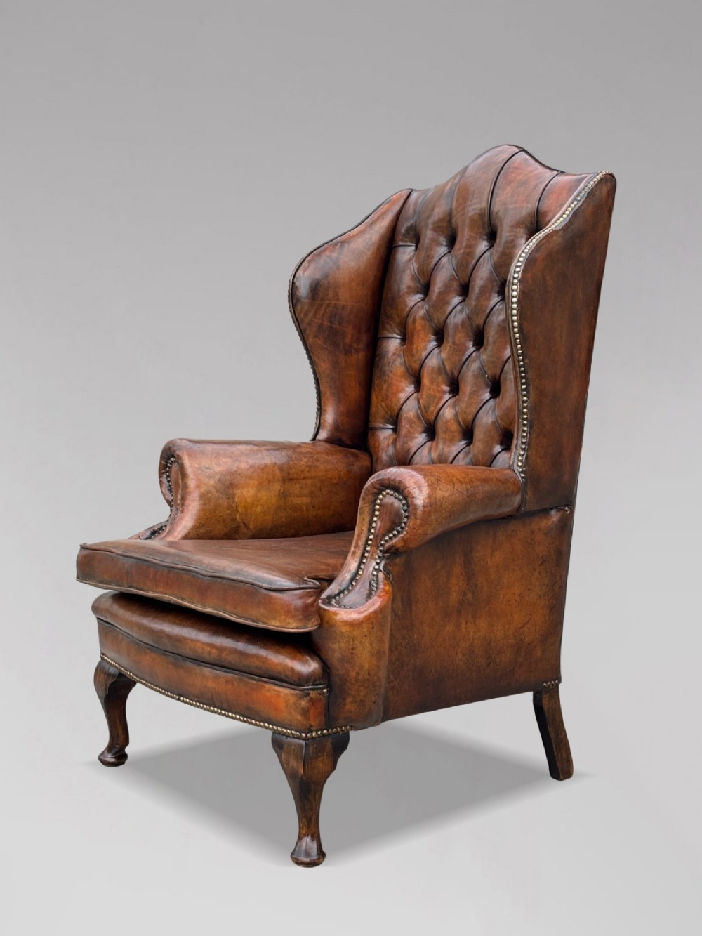 A 19th-century Victorian wingback armchair upholstered in deep-buttoned warm brown leather with brass stud detailing and carved mahogany legs. View IX