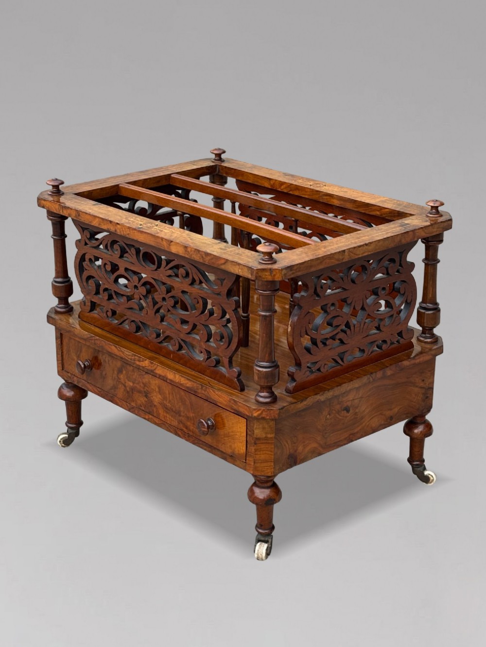 A mid-19th-century Victorian walnut music canterbury featuring open fretwork dividers, turned corner columns, a single base drawer, and turned legs with ceramic castors.