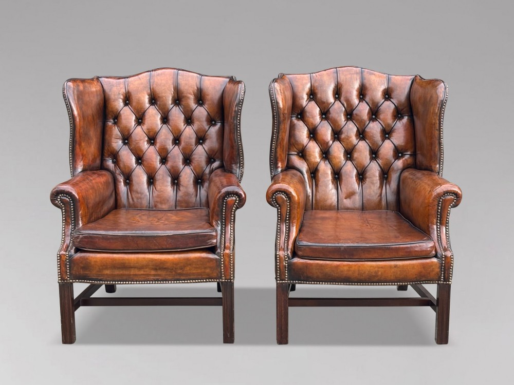 A pair of late Victorian wingback armchairs upholstered in deep-buttoned tan brown leather with brass stud trimming and dark wooden legs.