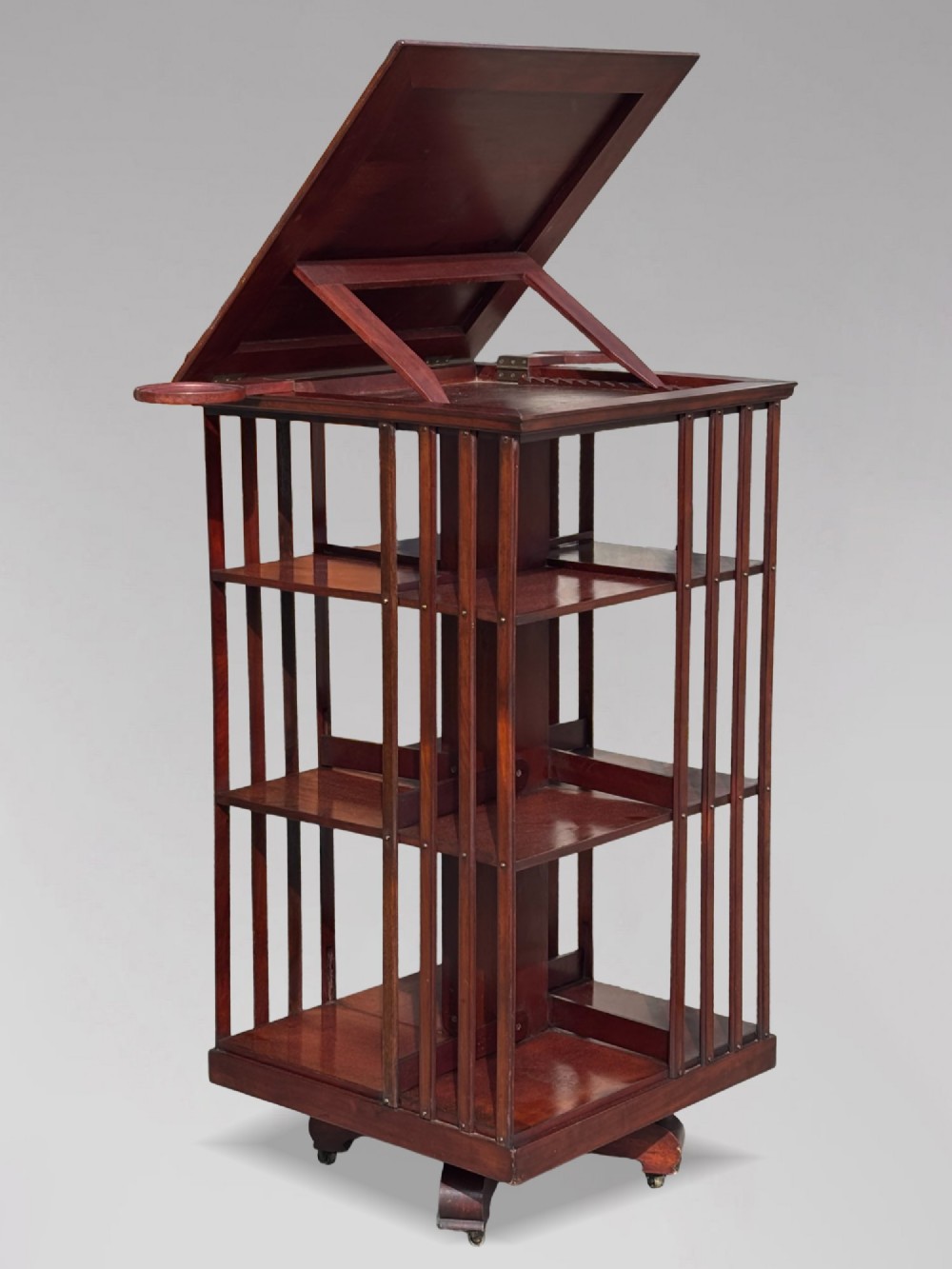 A tall nineteenth-century mahogany revolving bookcase featuring open slatted sides, tiered shelving, pull-out circular trays, and an angled reading slope with a burgundy leather surface. View III