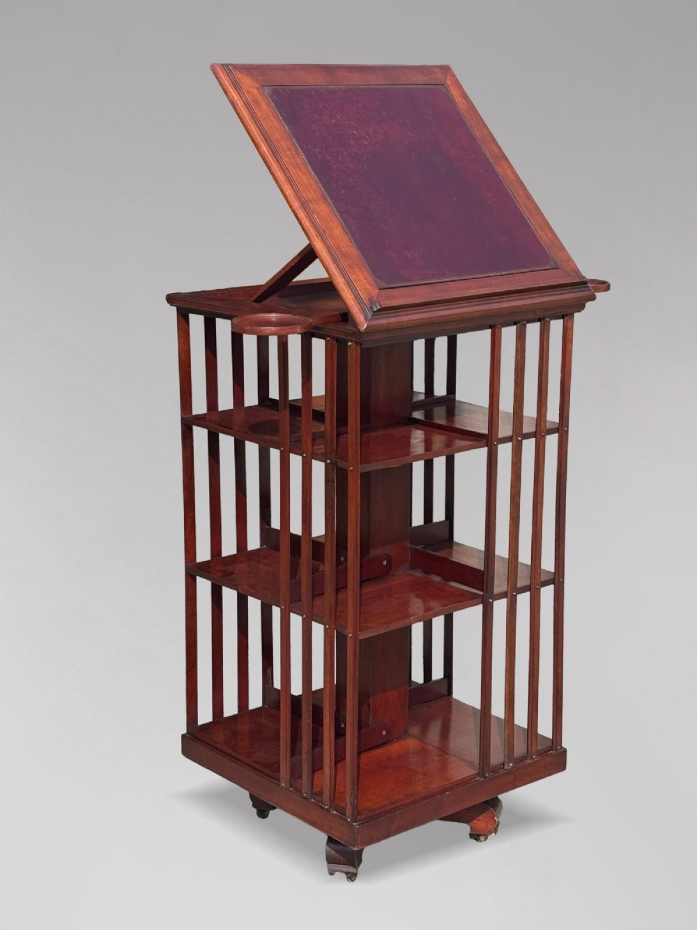 A tall nineteenth-century mahogany revolving bookcase featuring open slatted sides, tiered shelving, pull-out circular trays, and an angled reading slope with a burgundy leather surface. View V