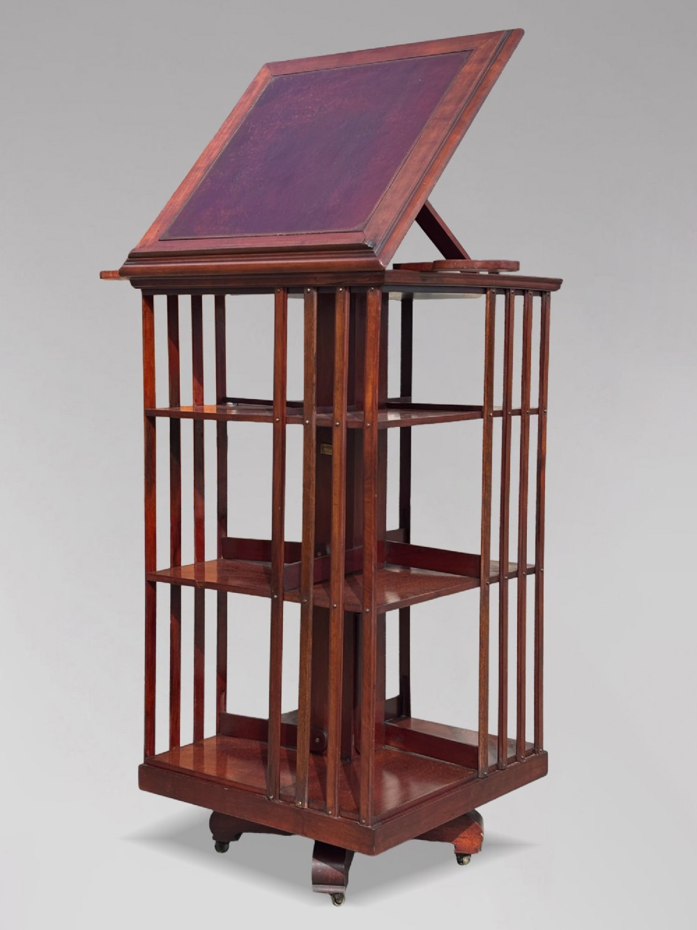 A tall nineteenth-century mahogany revolving bookcase featuring open slatted sides, tiered shelving, pull-out circular trays, and an angled reading slope with a burgundy leather surface. View X